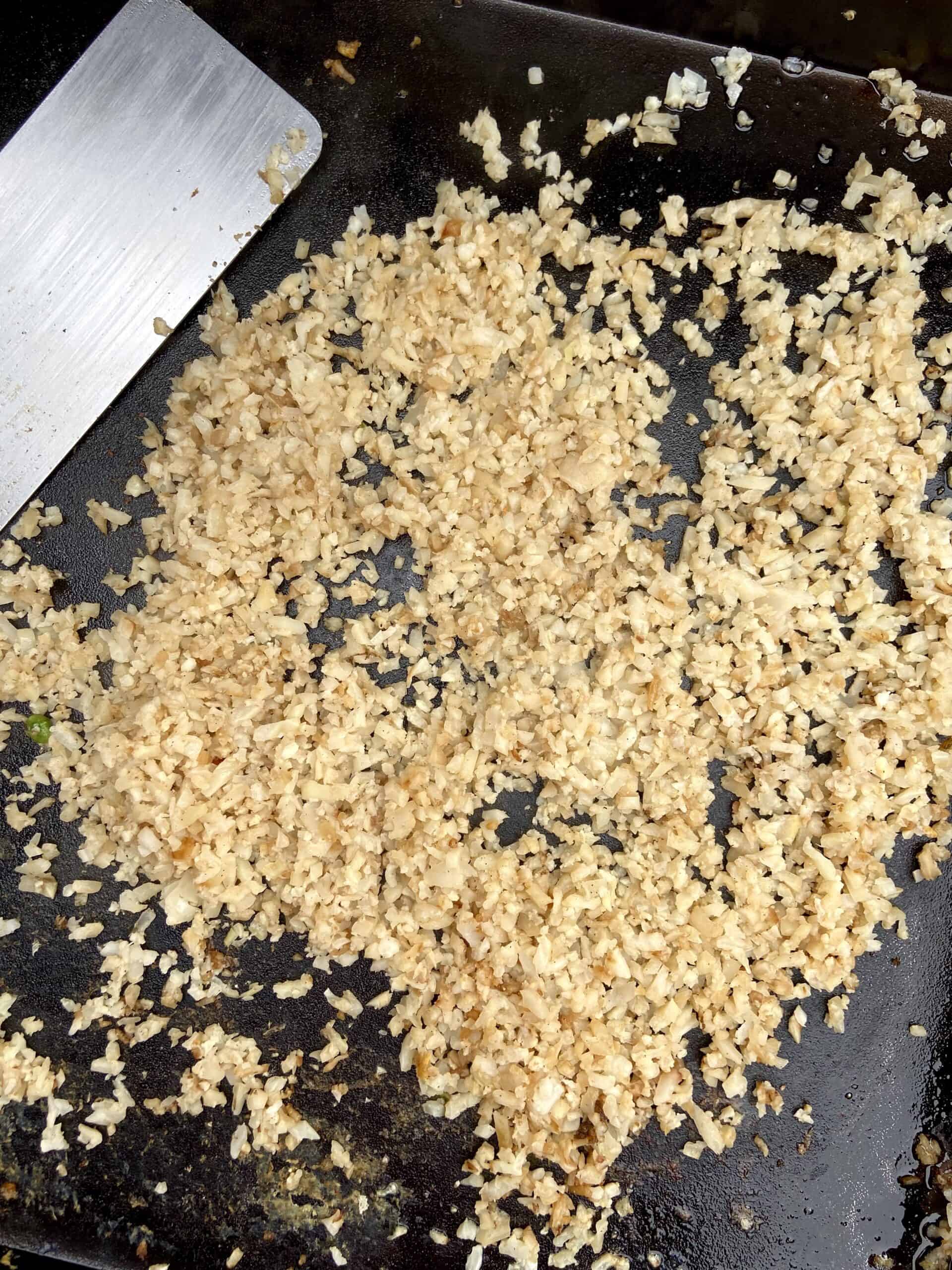 Cauliflower rice being cooked on a Blackstone griddle with a metal spatula near it.