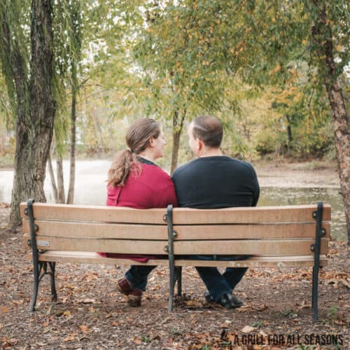 A man and woman sit on a park bench facing each other. They're near some trees and the season is fall.