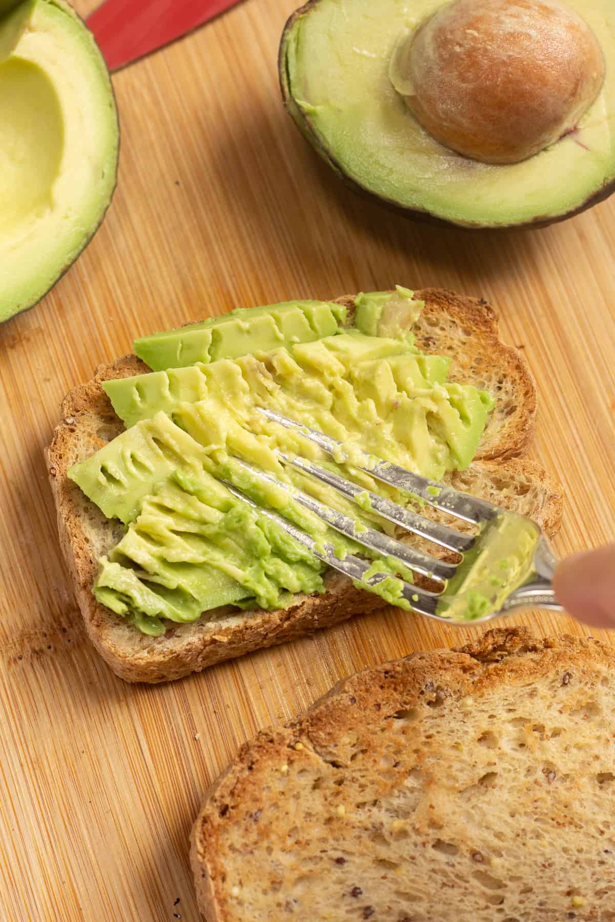 A fork smashing sliced avocado onto a piece of bread.
