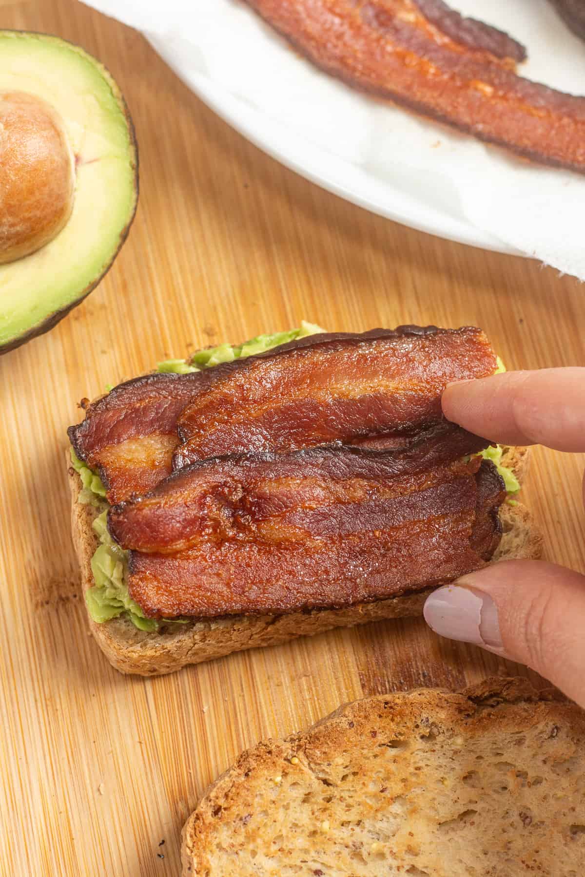 Placing slices of bacon on top of a piece of bread with avocado on it.