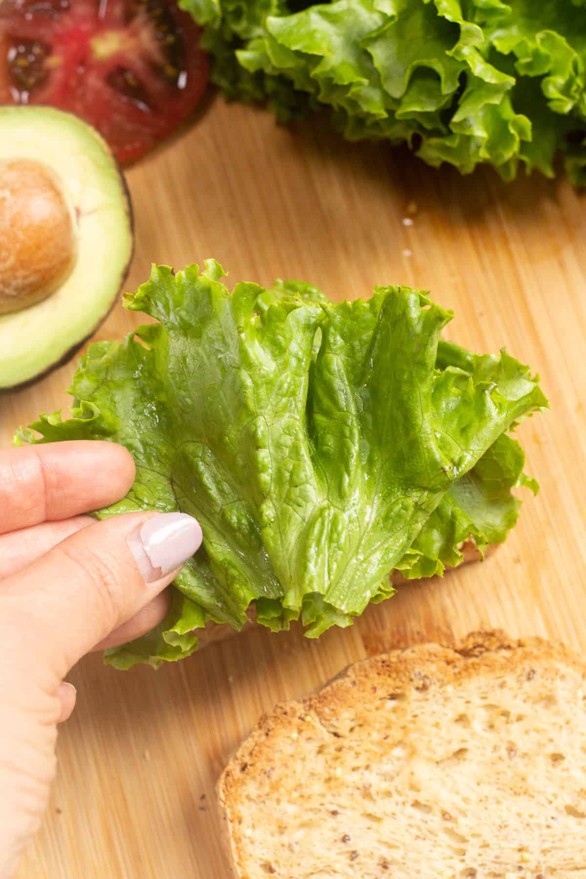 A hand placing a piece of lettuce on top of a blta sandwich.