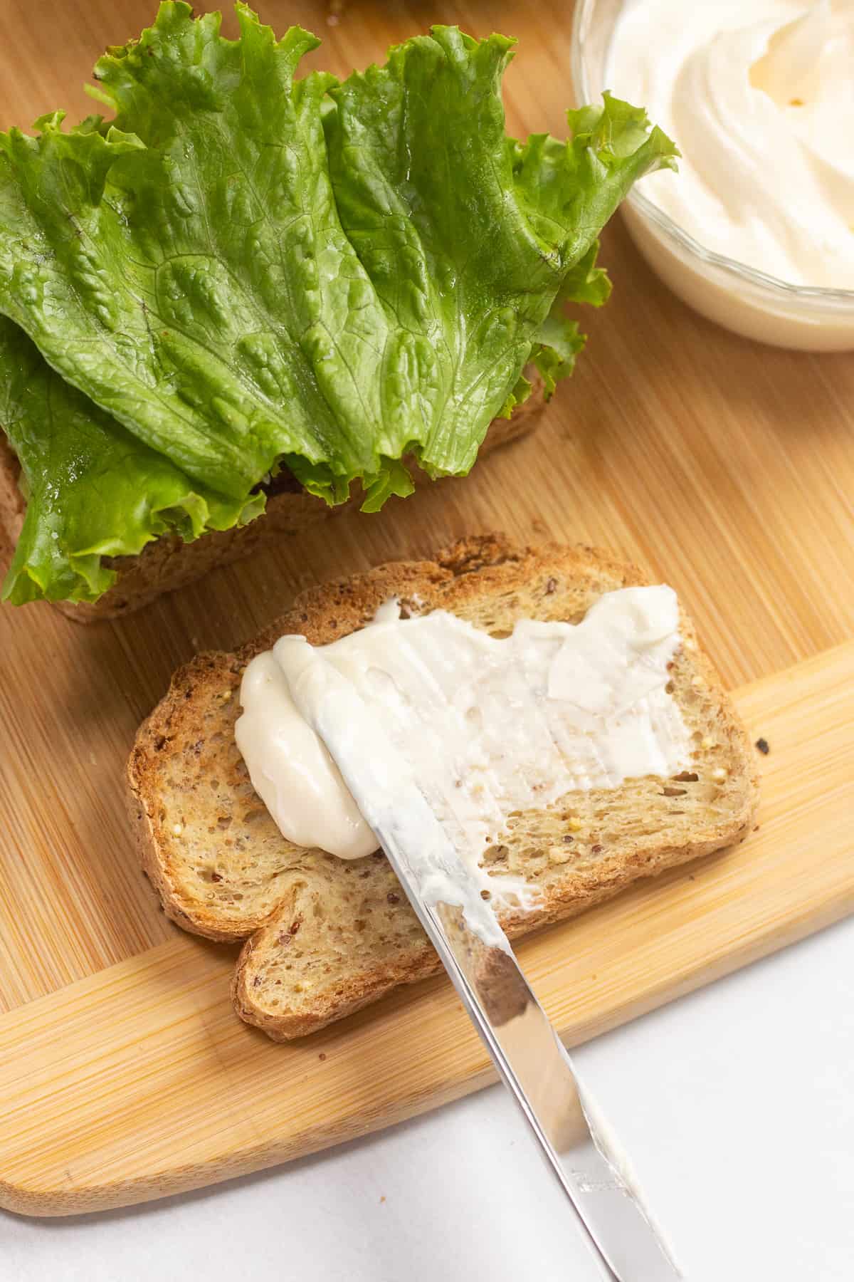 A table knife spreading mayo onto a toasted piece of bread on top of a wood cutting board.
