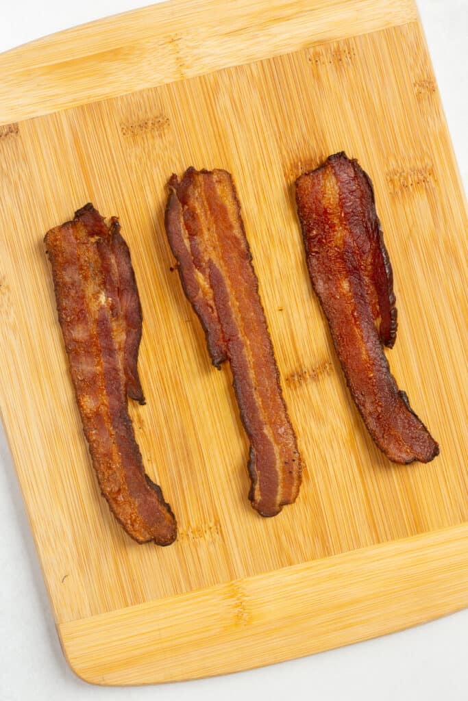 Three strips of cooked bacon on a wood cutting board.