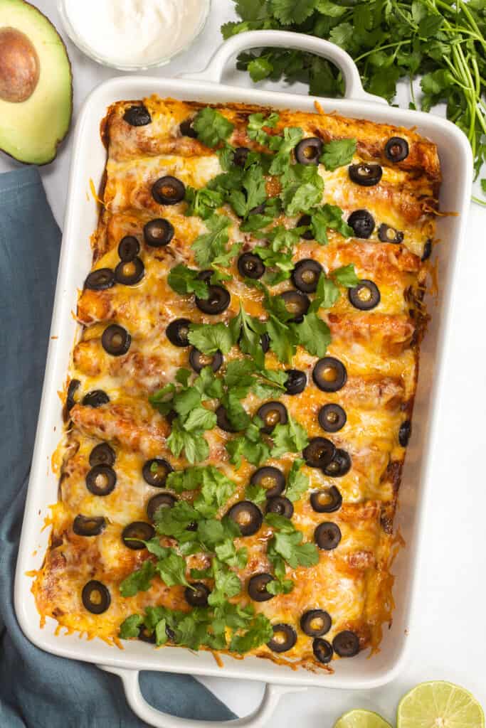 a top down shot of an enchilada casserole in a white dish with sliced olives and cilantro on top.