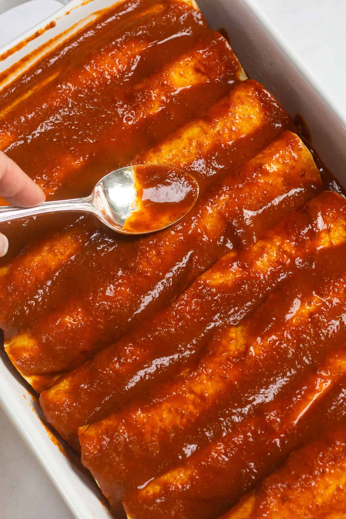 A spoon spreading enchilada sauce over rolled tortillas in a casserole dish.