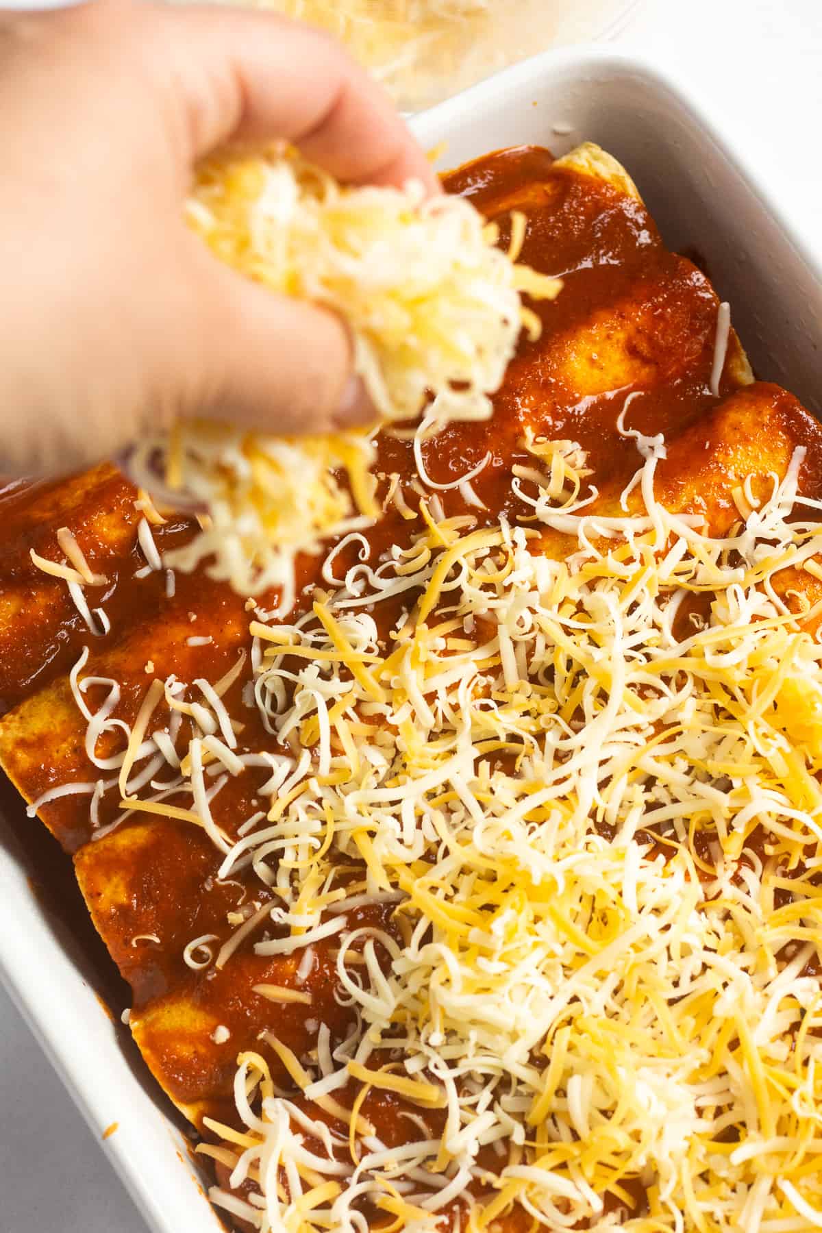 A hand sprinkling cheese over sauced rolled tortillas in a casserole dish.