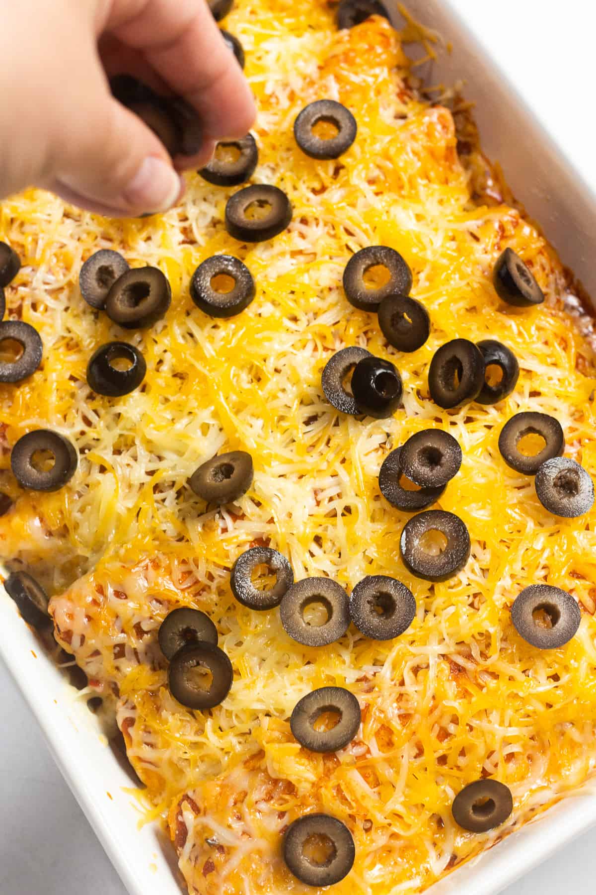 A hand sprinkling sliced olives over half cooked enchiladas in a white baking dish.