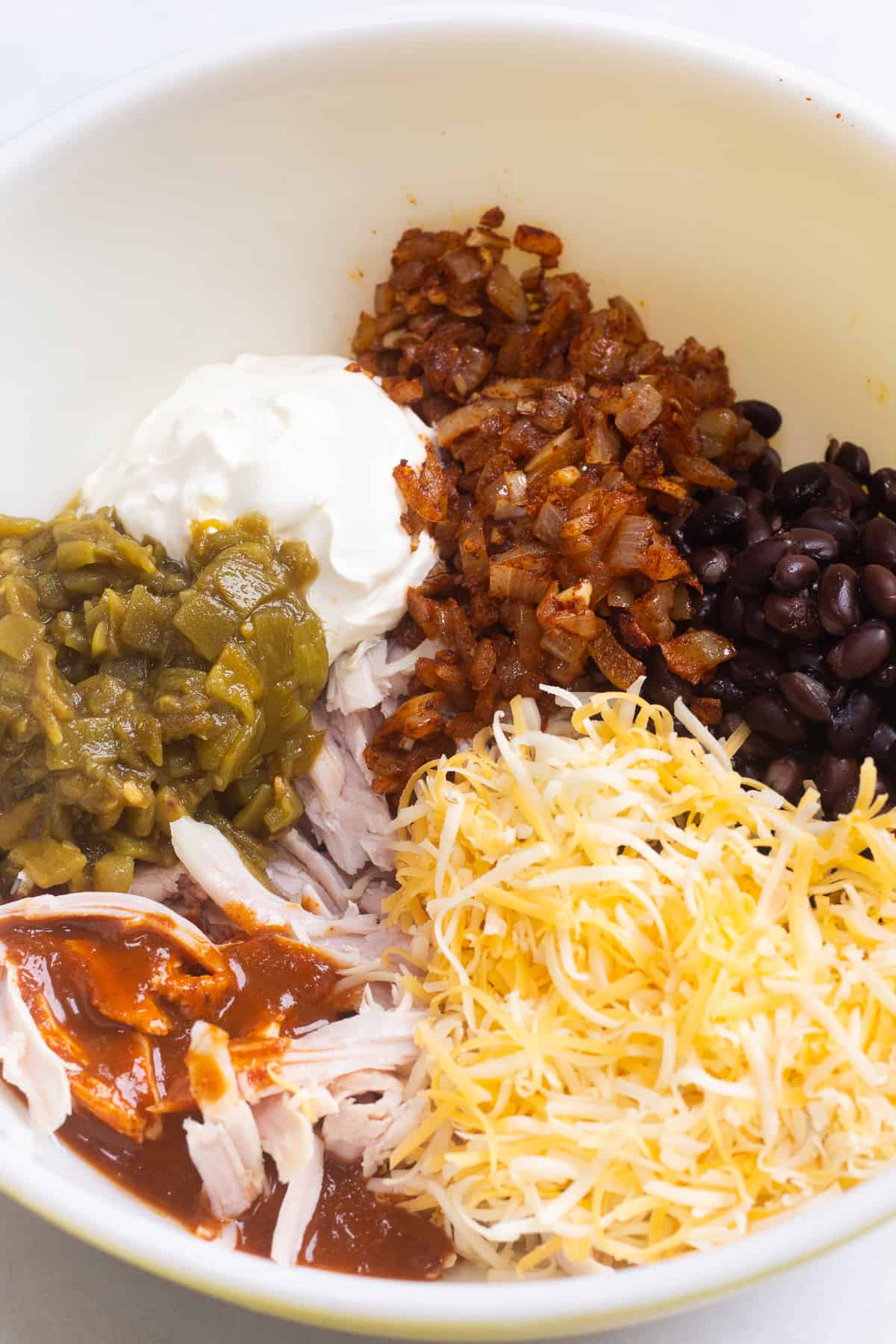 A large bowl with shredded cheese, chiles, sour cream, black beans, shredded turkey, and enchilada sauce in it.