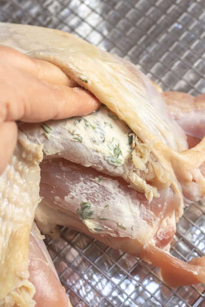 a hand spreading herb butter onto a raw turkey breast.