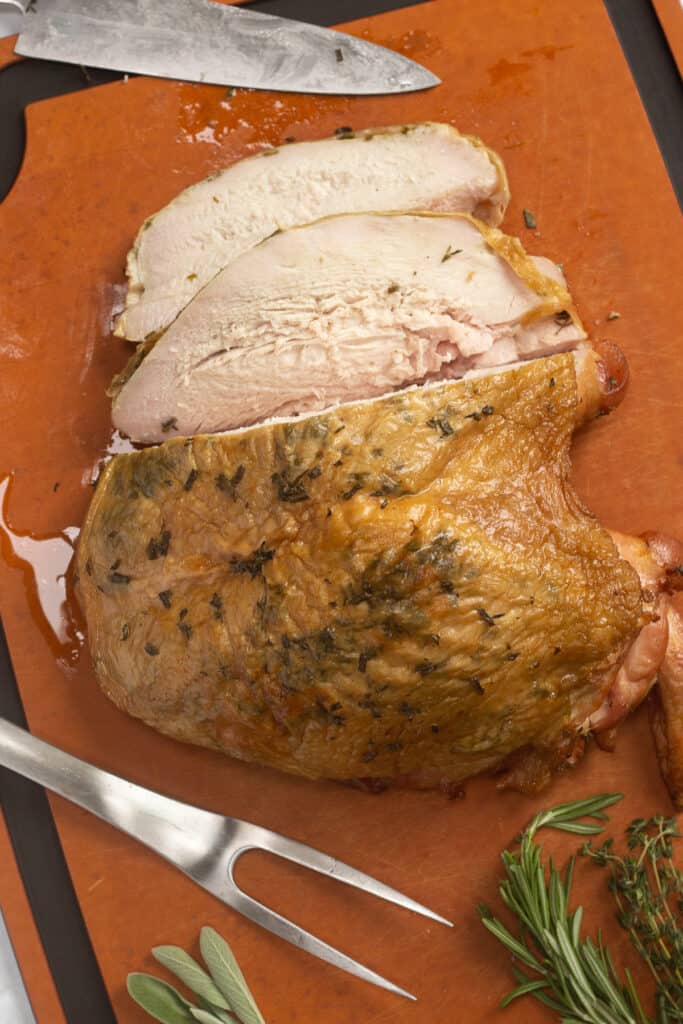 a top down shot of a Traeger smoked turkey breast on a wood cutting board with herbs and a knife next to it.