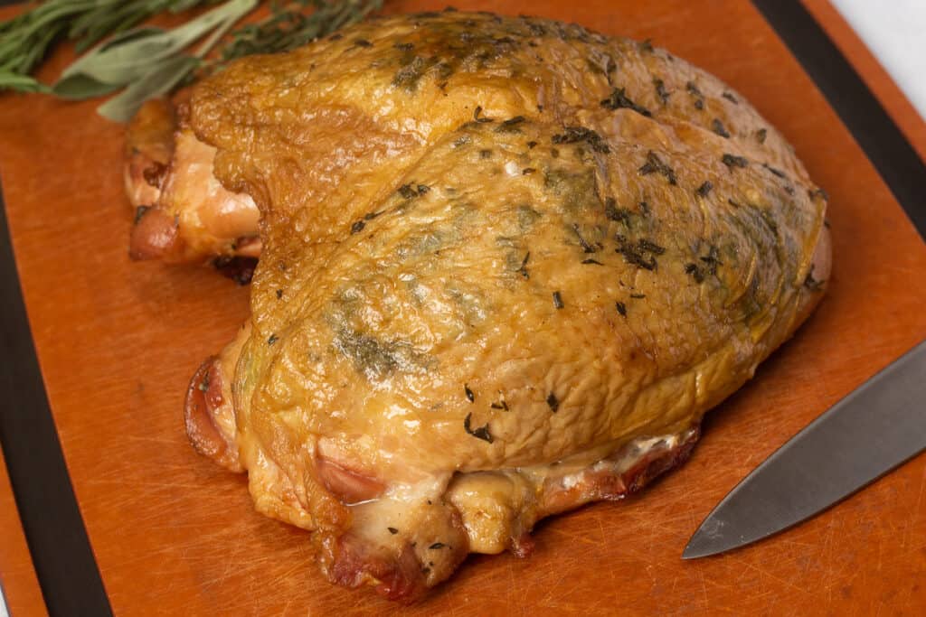 a smoked turkey breast resting on a wood cutting board with a knife and herbs next to it.
