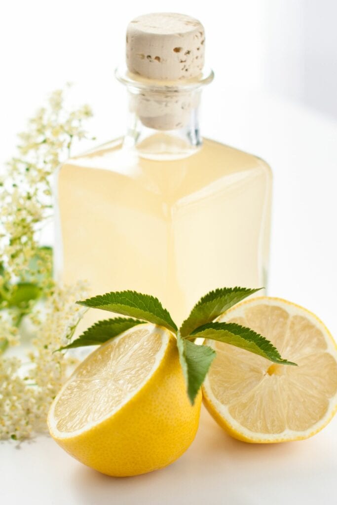 A bottle with lemon syrup in it with a split lemon and some flowers next to it.