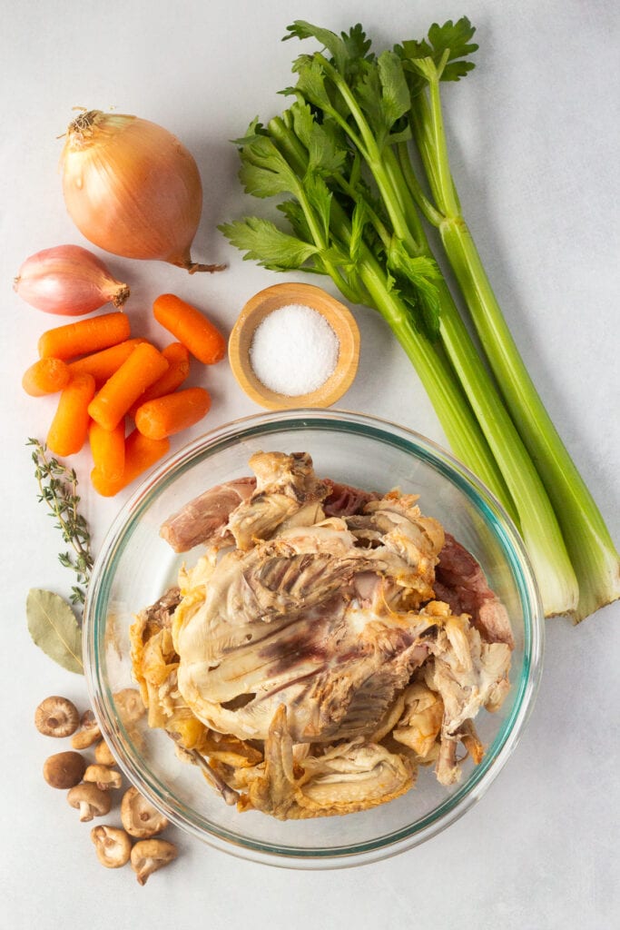 A top down shot of ingredients for chicken stock: celery, carrots, mushrooms, salt, onion, a shallot, bay leaf, thyme, and a chicken carcass in a bowl.