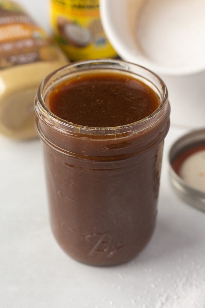 A close up of small mason jar with a brown-colored marinade next to bottles, salt, and a lid.