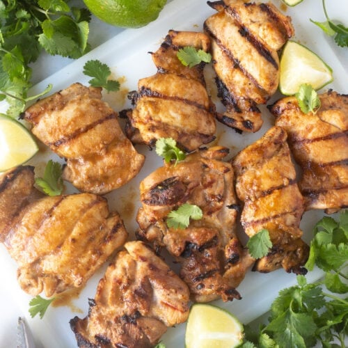 Top down shot of Mexican grilled chicken thighs on a white cutting board with limes and cilantro scattered about.