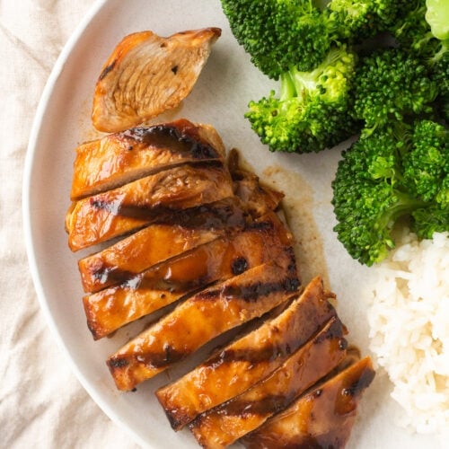 Top down shot of a grilled and sliced boneless chicken breast with brown sauce on a white plate next to cooked rice and broccoli.