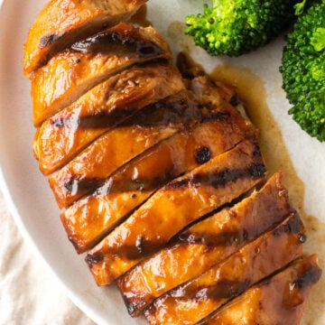 Top down shot of sliced marinated and grilled boneless skinless chicken breast on a white plate next to cooked broccoli.