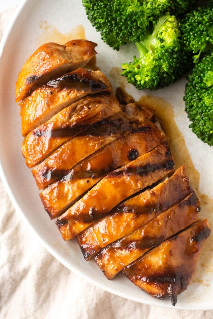 Top down shot of sliced marinated and grilled boneless skinless chicken breast on a white plate next to cooked broccoli.