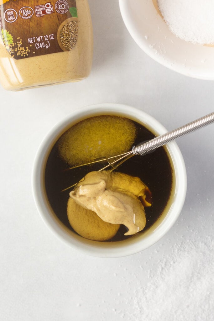 A brown sauce with dijon mustard and sauce in  a small white bowl with a whisk in it.