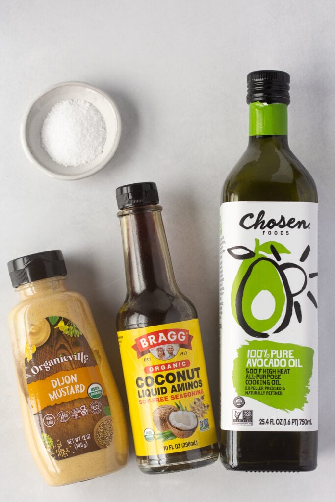 A dijon mustard bottle, a coconut aminos bottle, an avocado oil bottle, and a small white bowl with kosher salt in it.