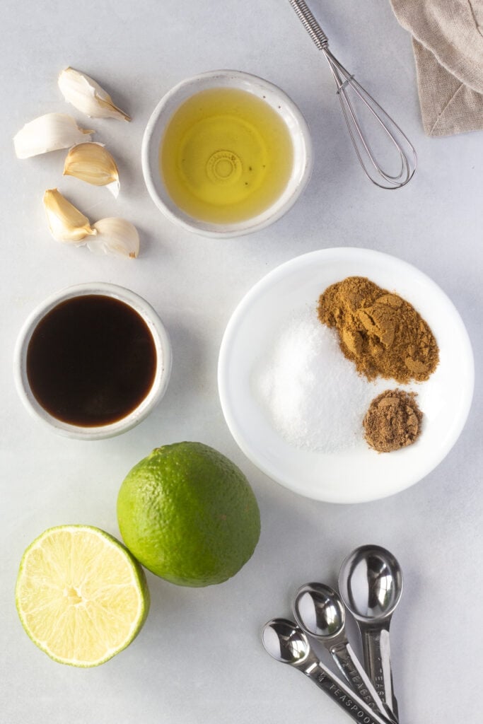 A top down shot of ingredients for Mexican chicken marinade in small white bowls, including oil, spices, limes, and garlic cloves.