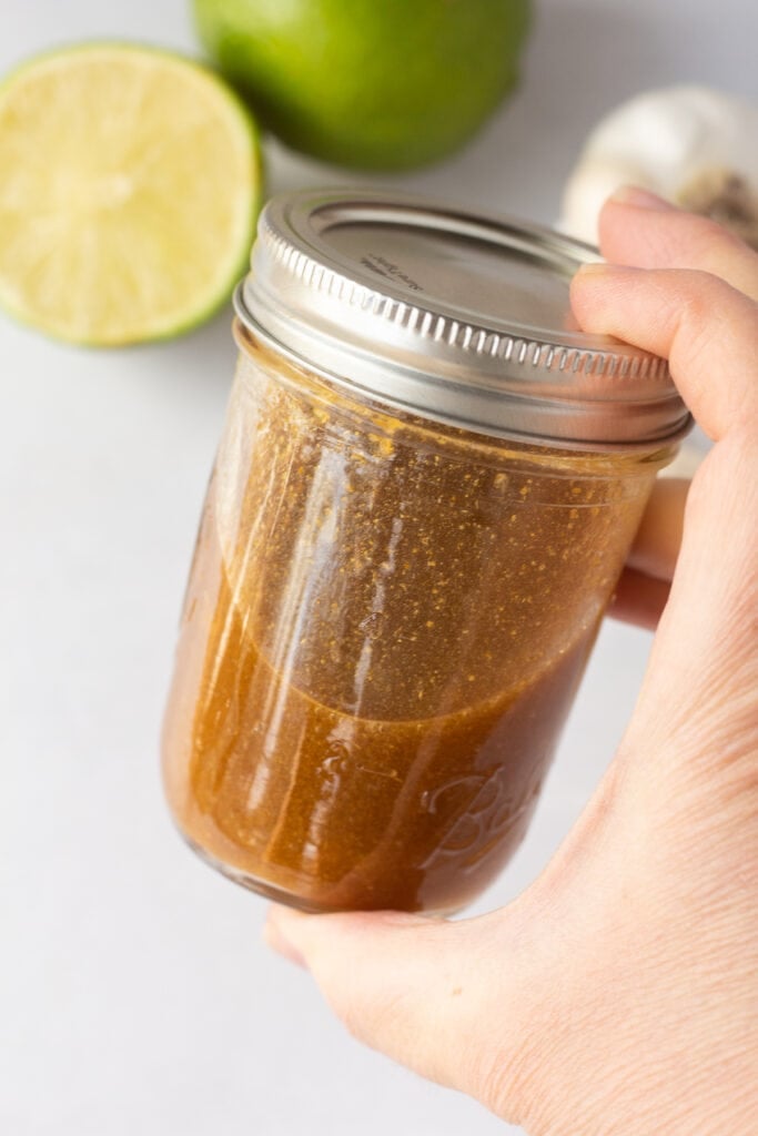 A hand holding a small lidded mason jar with a brown chicken marinade in it.