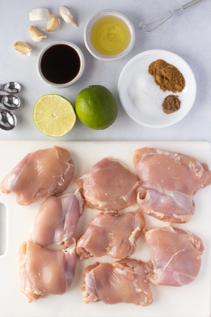 top down shot of ingredients for grilled Mexican chicken thighs, including raw boneless skinless chicken thighs, lime, oil, spices, garlic, and coconut aminos.