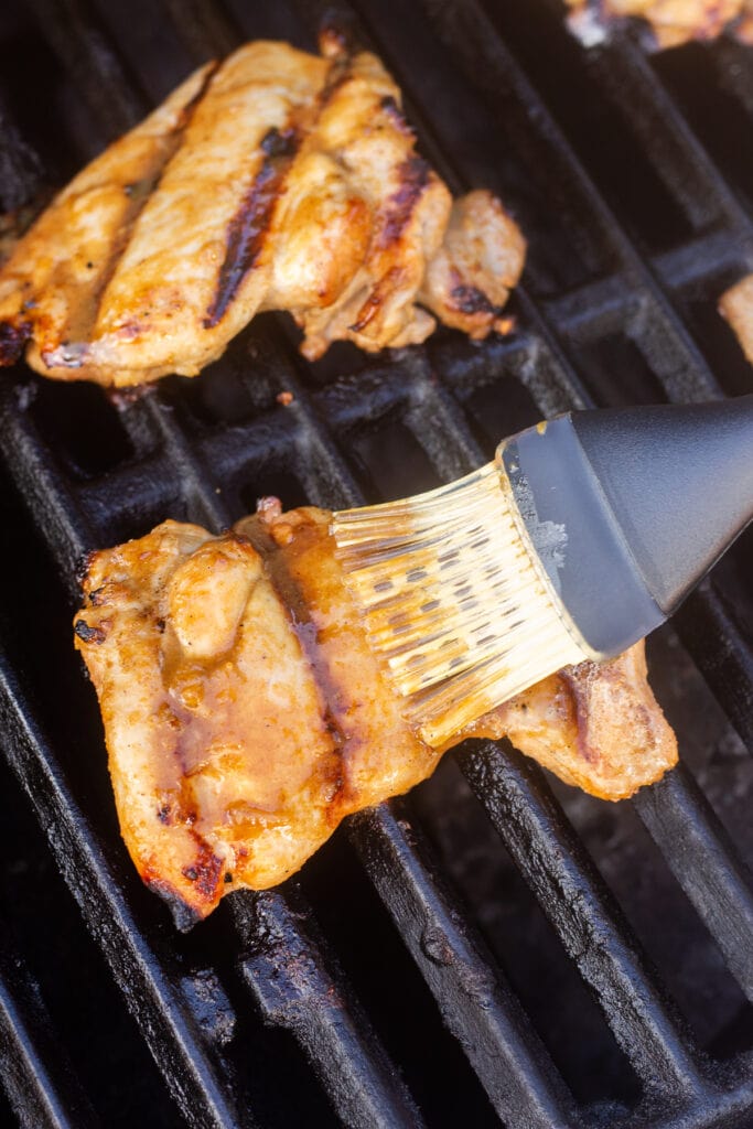A basting brush putting marinade onto a boneless grilled chicken thigh on the grill.