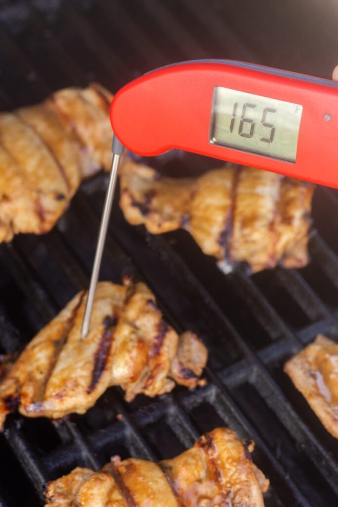 A meat thermometer measuring the internal temperature of grilled boneless chicken thighs on a grill.