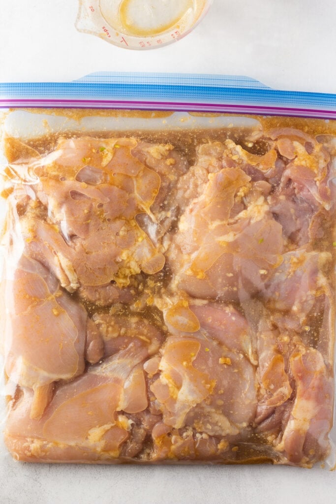 raw chicken thighs marinating in a brown liquid inside a large ziploc plastic bag.