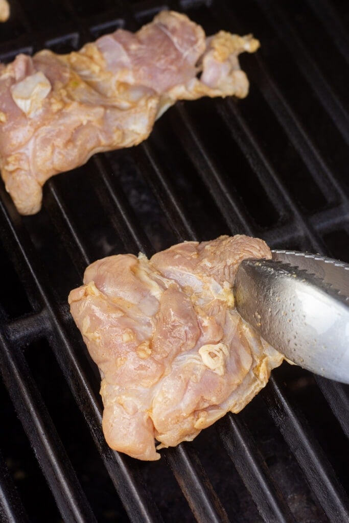 Placing a raw marinated boneless skinless chicken thigh on the grill with tongs.
