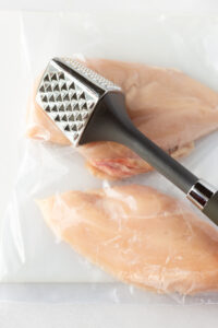 A meat mallet pounding raw chicken breasts that are in a plastic bag on top of a white cutting board.