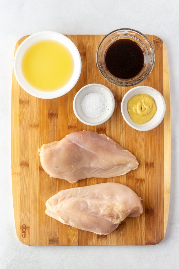 Top down shot of two raw boneless skinless chicken breasts on a cutting board with small bowls containing marinade ingredients.