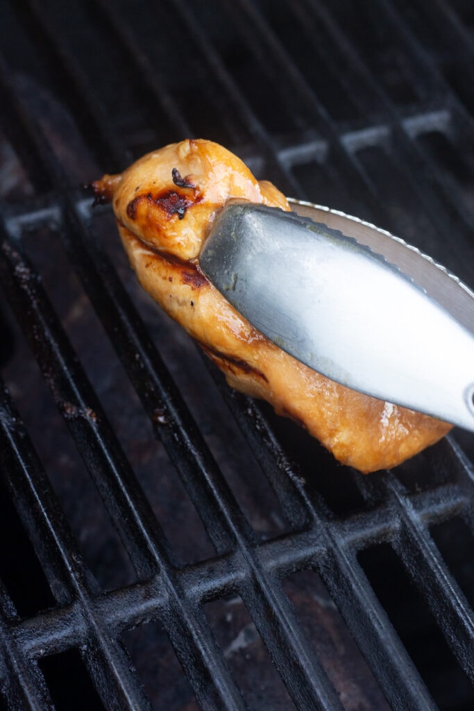 A pair of tongs flipping a boneless chicken breast over on a grill.