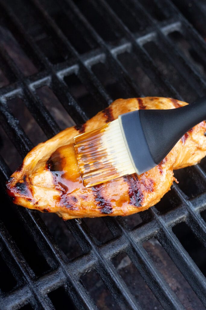 A basting brush brushing a glaze on a boneless grilled chicken breast on a grill.