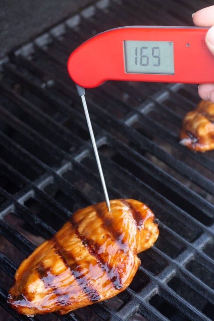 A meat thermometer measuring the internal temp of a chicken breast on a grill. The temperature reads 165 degrees F