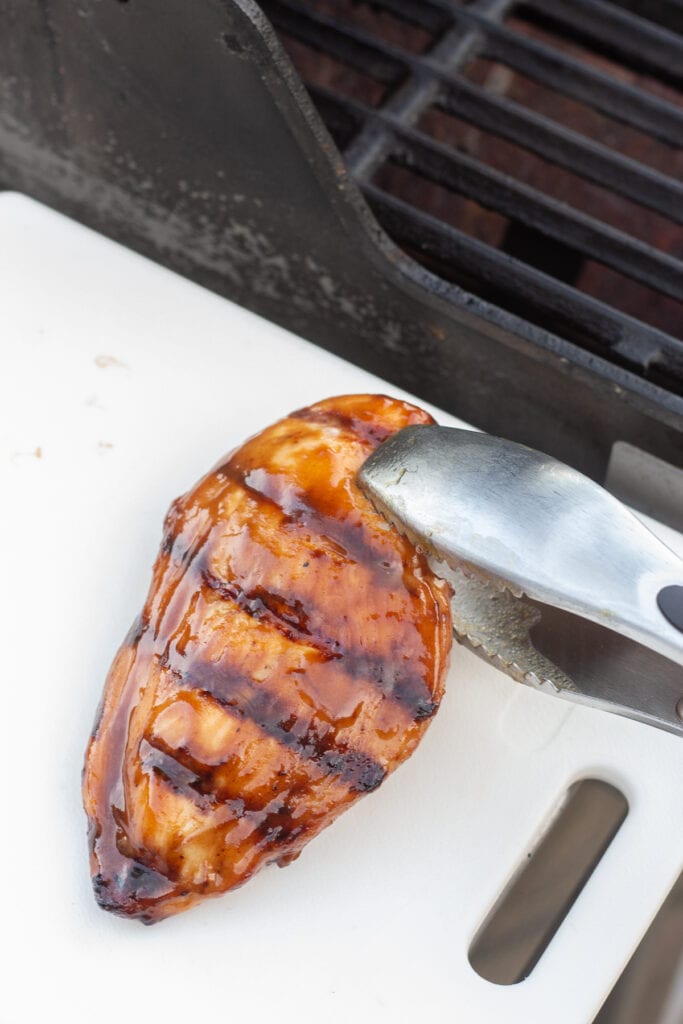 A pair of tongs removing grilled boneless chicken breast from the grill onto a white cutting board.