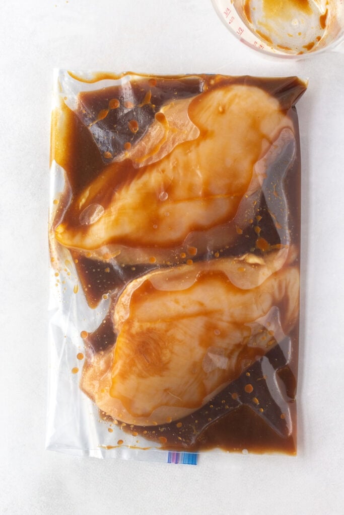 Top down shot of raw boneless skinless chicken breasts in a brown marinade in a ziploc bag.
