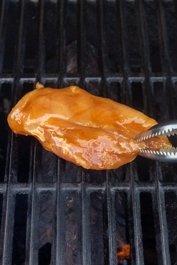 Placing a raw marinated boneless chicken breast on a hot grill with tongs.