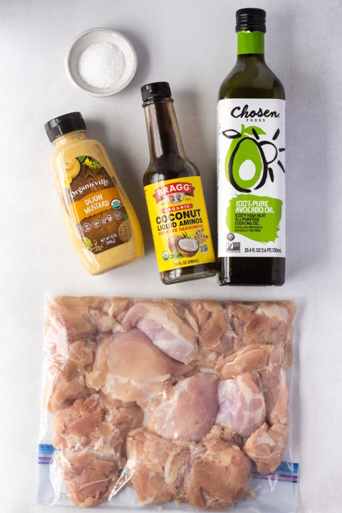 Top down shot of boneless skinless raw chicken thighs in a ziploc bag next to bottles of dijon mustard, coconut aminos, and avocado oil. A white dish with kosher salt is in the upper left corner.
