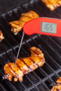 An instant read thermometer measuring the internal temp of chicken thighs on the grill.