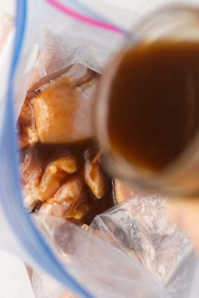 pouring brown marinade from a mason jar into a ziploc bag with raw boneless skinless chicken thighs in it.