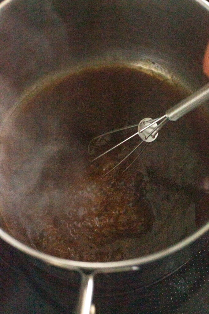 A close up of a brown chicken marinade being boiled in a small sauce pan with a whisk.
