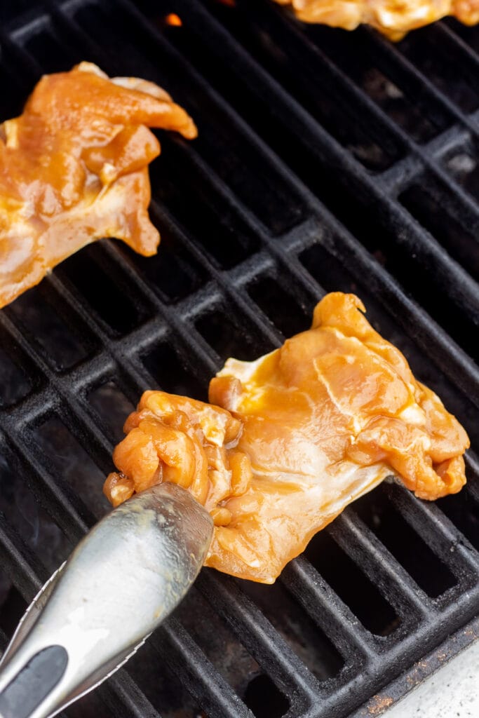A pair of tongs placing raw chicken thighs on a hot grill.