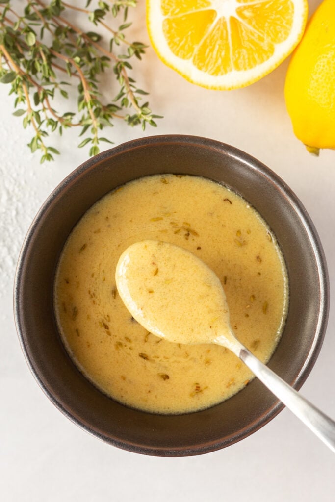 top down shot of a bowl with a dijon mustard marinade and a spoon holding some of the marinade above the bowl. Some fresh thyme and cut lemon surround the bowl.