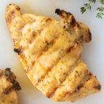 Top down shot of a grilled dijon chicken breast on a white cutting board with cut lemons and fresh thyme surrounding it.