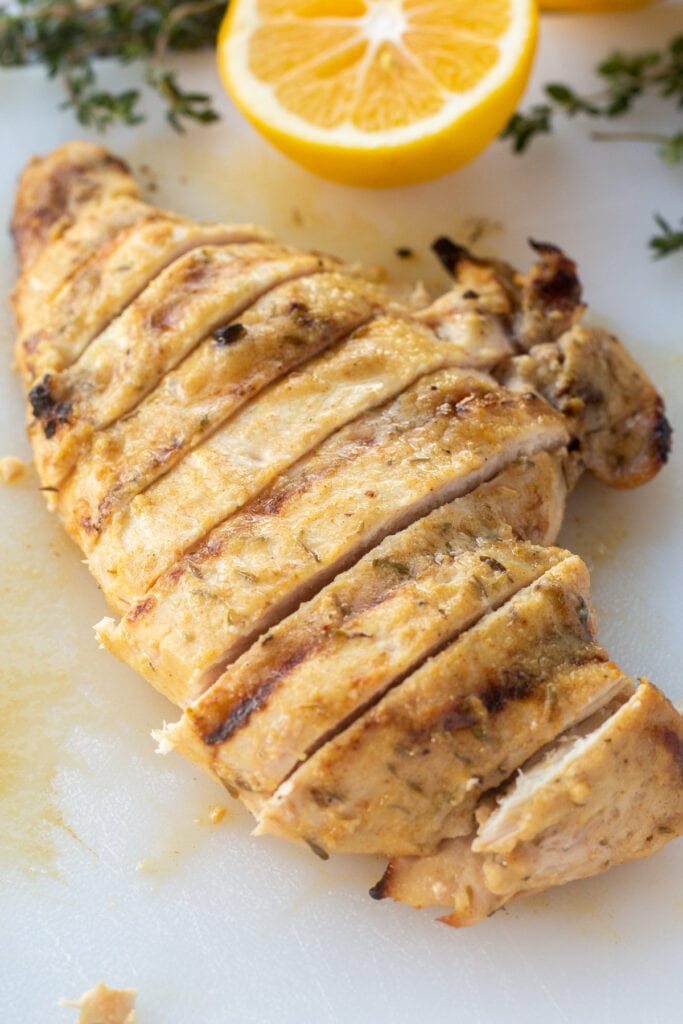 Close up of sliced grilled dijon chicken breast on a white cutting board with lemon and fresh thyme around it.