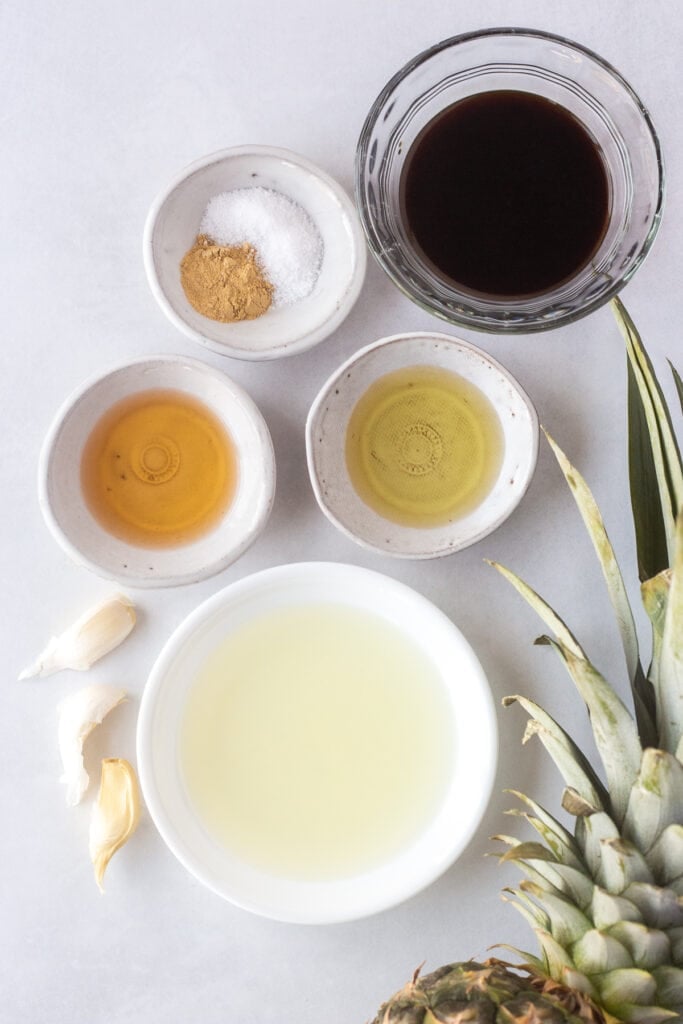 Top down shot of small white and clear bowls with different liquids and spices in them, with 3 garlic cloves to the left and a fresh pineapple peeking out on the right side.