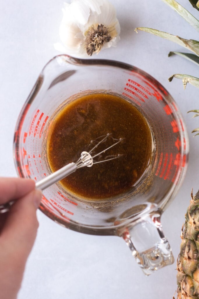 A hand whisking brown marinade ingredients in a measuring cup on a white background.