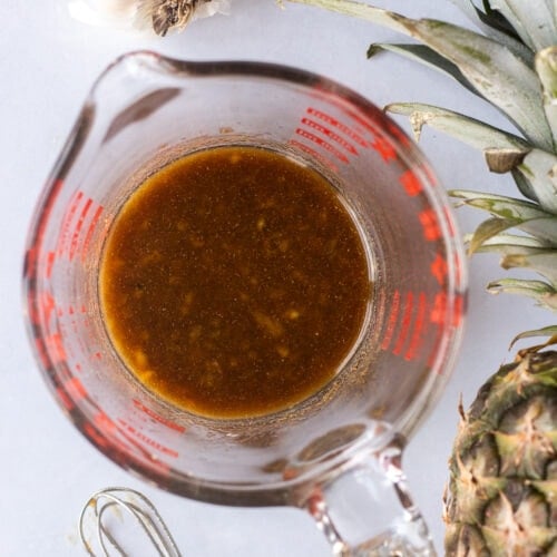 Top down shot of a measuring cup with a brown marinade in it. A garlic head, a pineapple, and a small whisk are off to different sides of the cup.